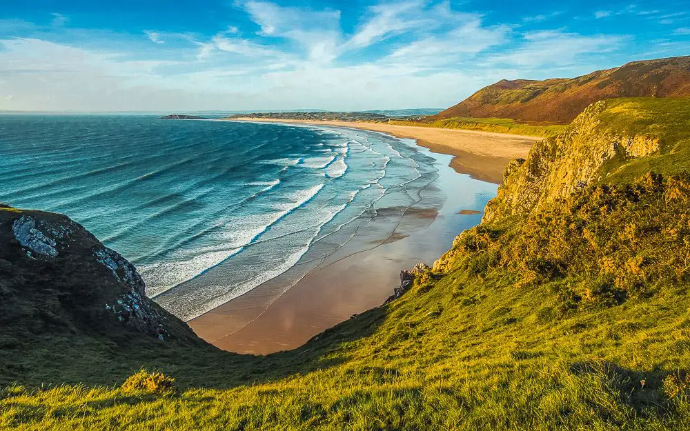 Türkisblaues Meer, grüne Wiesen und ein Sandstrand an der Küste von Wales an einem sonnigen Tag mit blauen Himmel. 